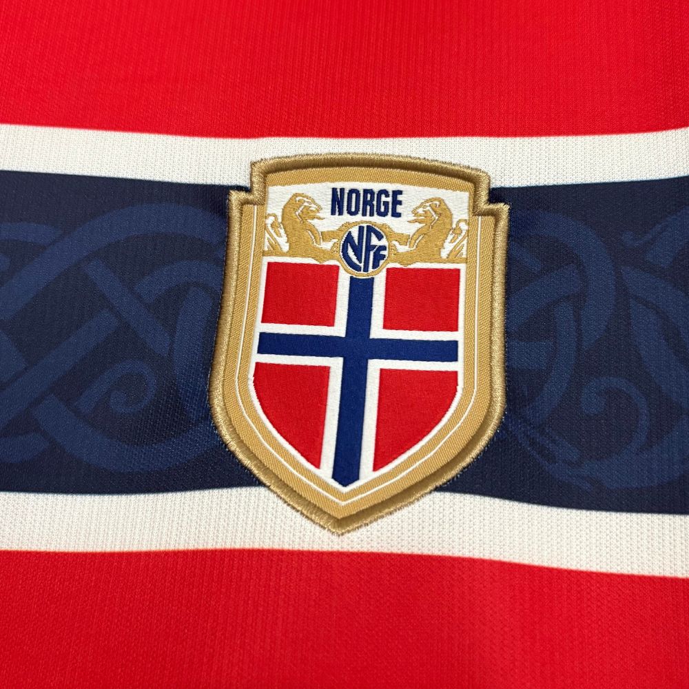 Camisa Nike Noruega 26/27 Home, , PIQUE DE JOGADOR, A Camisa Noruega 2026 Home representa o uniforme principal da seleção da Noruega para o ciclo da Copa do Mundo de 2026, trazendo um design moderno com forte inspiração na identidade nacional do país. O modelo apresenta a tradicional base em vermelho, cor histórica da seleção norueguesa, combinada com uma releitura marcante da bandeira do país. Um grande cruzamento em azul marinho com contornos brancos percorre o peito e o corpo da camisa, criando um visual