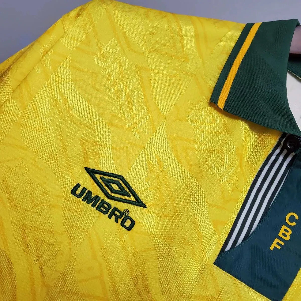 Camisa Umbro Retrô Seleção Brasil 1991/92 Home, , PIQUE DE JOGADOR, Reviva um período marcante da história da Seleção Brasileira com a Camisa Umbro Retrô Seleção Brasil 1991/92 Home. Inspirada no uniforme utilizado no início da década de 1990, essa peça resgata um design clássico que marcou a transição entre gerações do futebol brasileiro. O modelo apresenta o tradicional amarelo da Seleção com detalhes verdes, seguindo o estilo característico da Umbro na época, com linhas simples e identidade forte dentro 